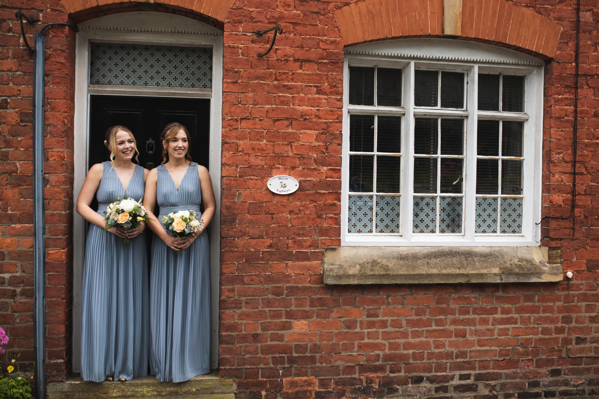 Two bridesmaids in blue dresses standing in a doorway of a red brick house