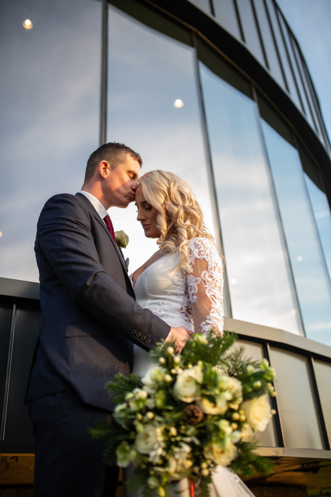 Groom kissing the bride's forehead against a glass building at golden hour