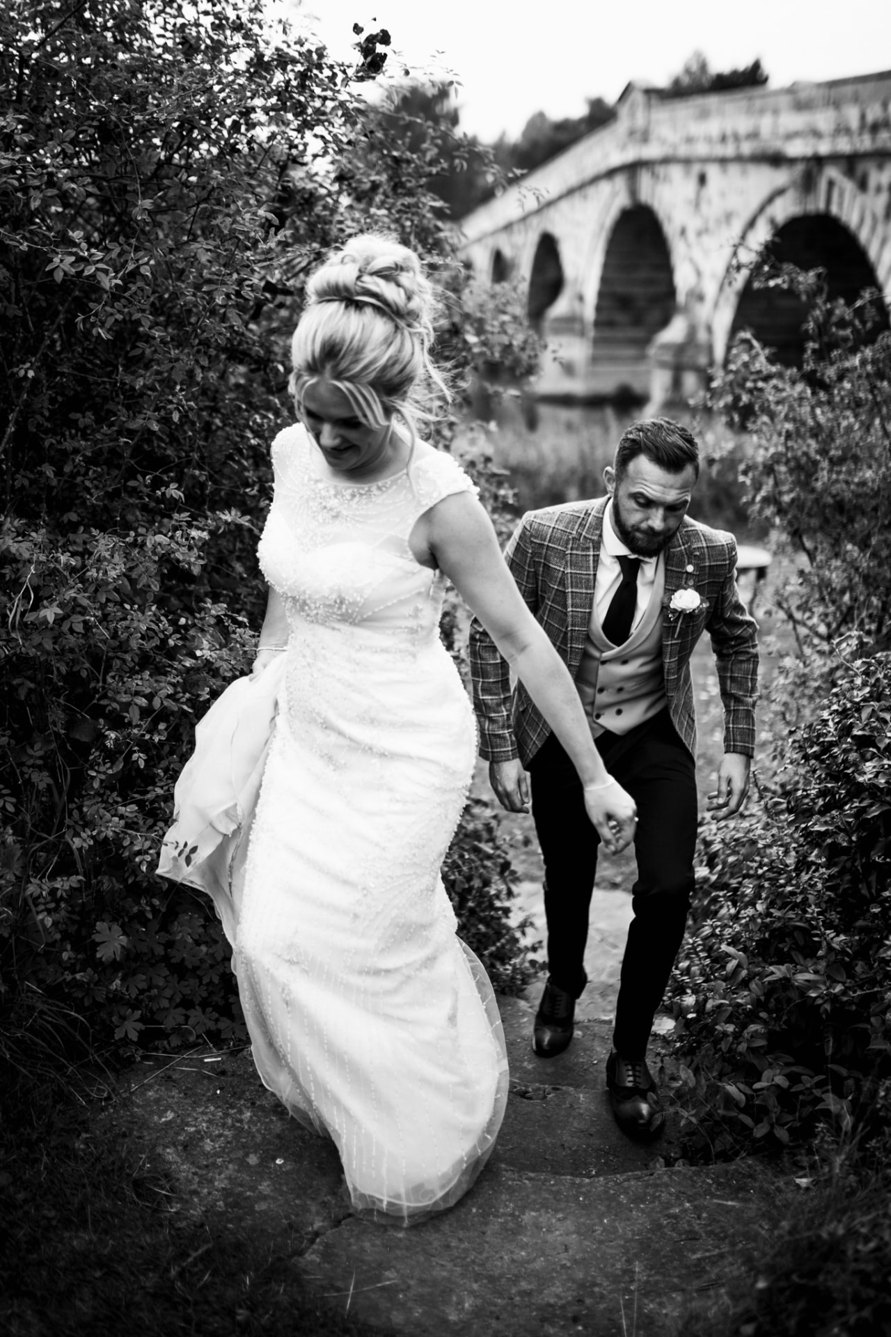 Bride leading the groom up stone steps by an old arched bridge, black and white