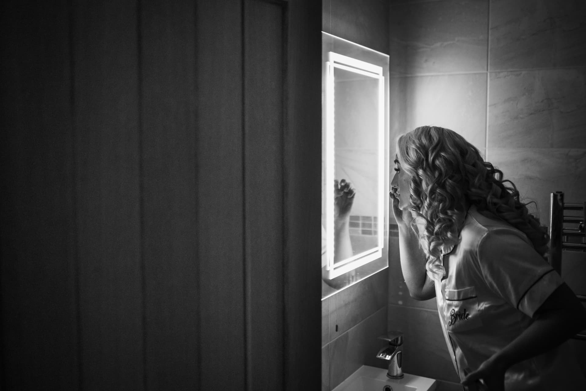 Bride in a robe embroidered with the word Bride, touching up her make-up in a glowing mirror, black and white