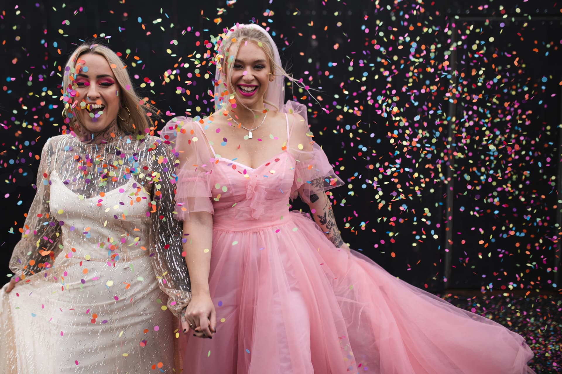 Two tattooed brides in silver and pink wedding dresses laughing in colourful confetti