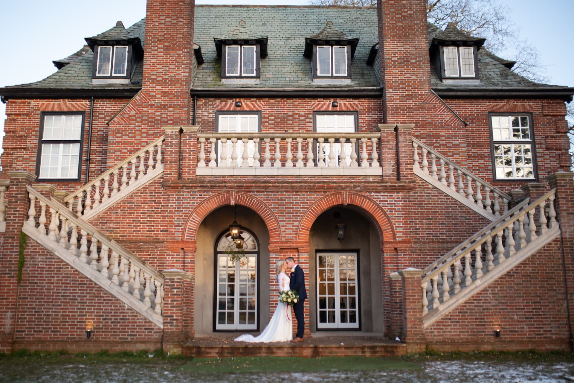 Bride and groom kissing between two brick archways in front of a red-brick manor house