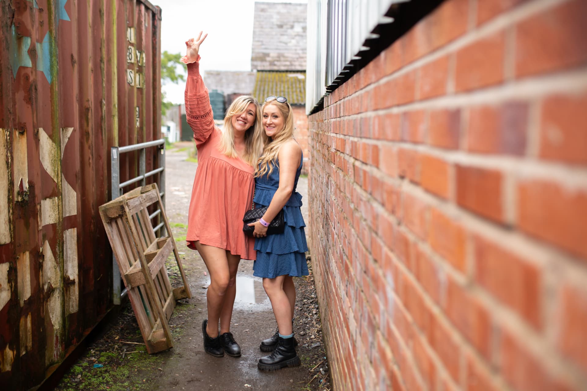Two guests in boots throwing a peace sign between a shipping container and brick wall