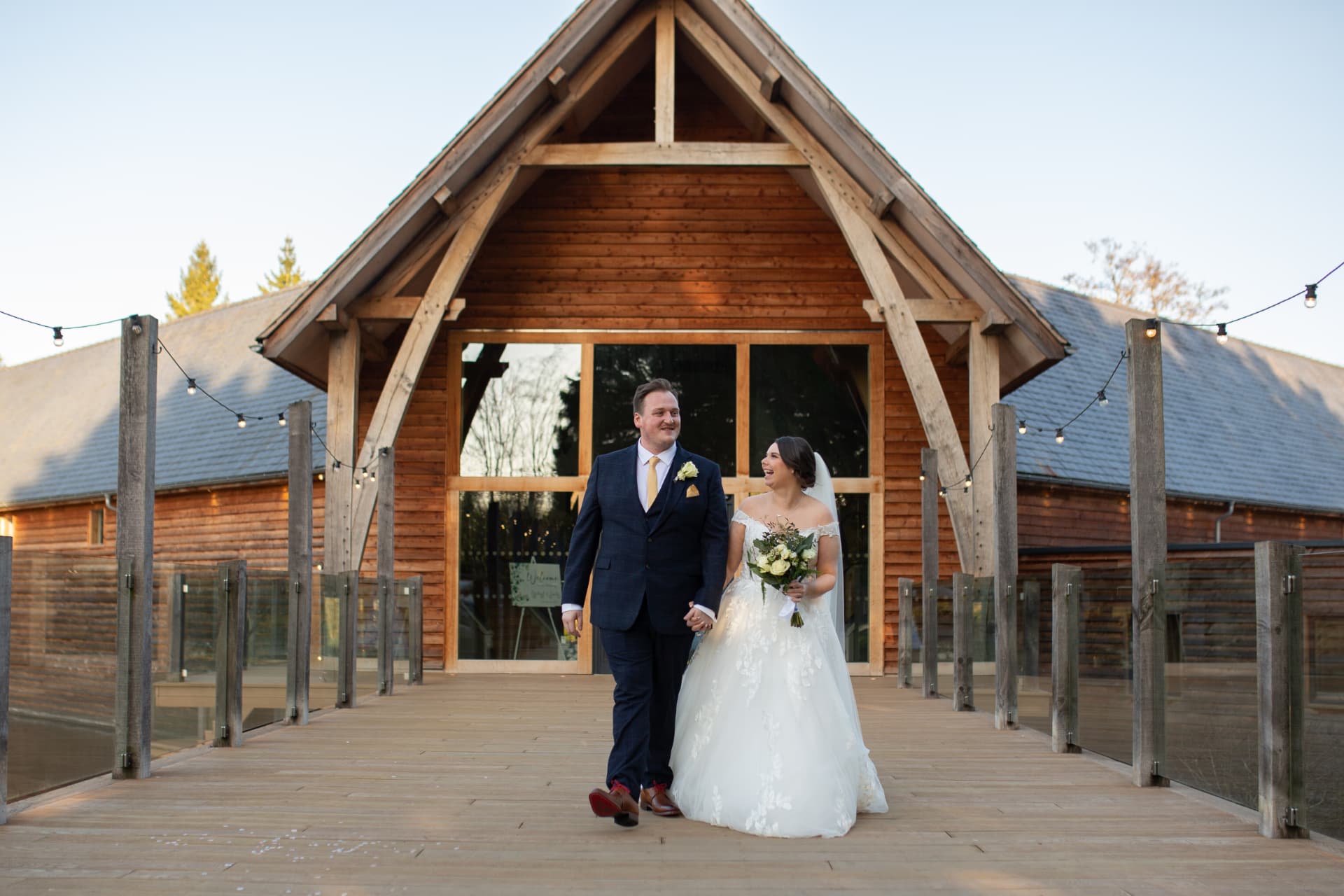 Bride and groom walking hand in hand along a wooden walkway outside a timber-framed barn