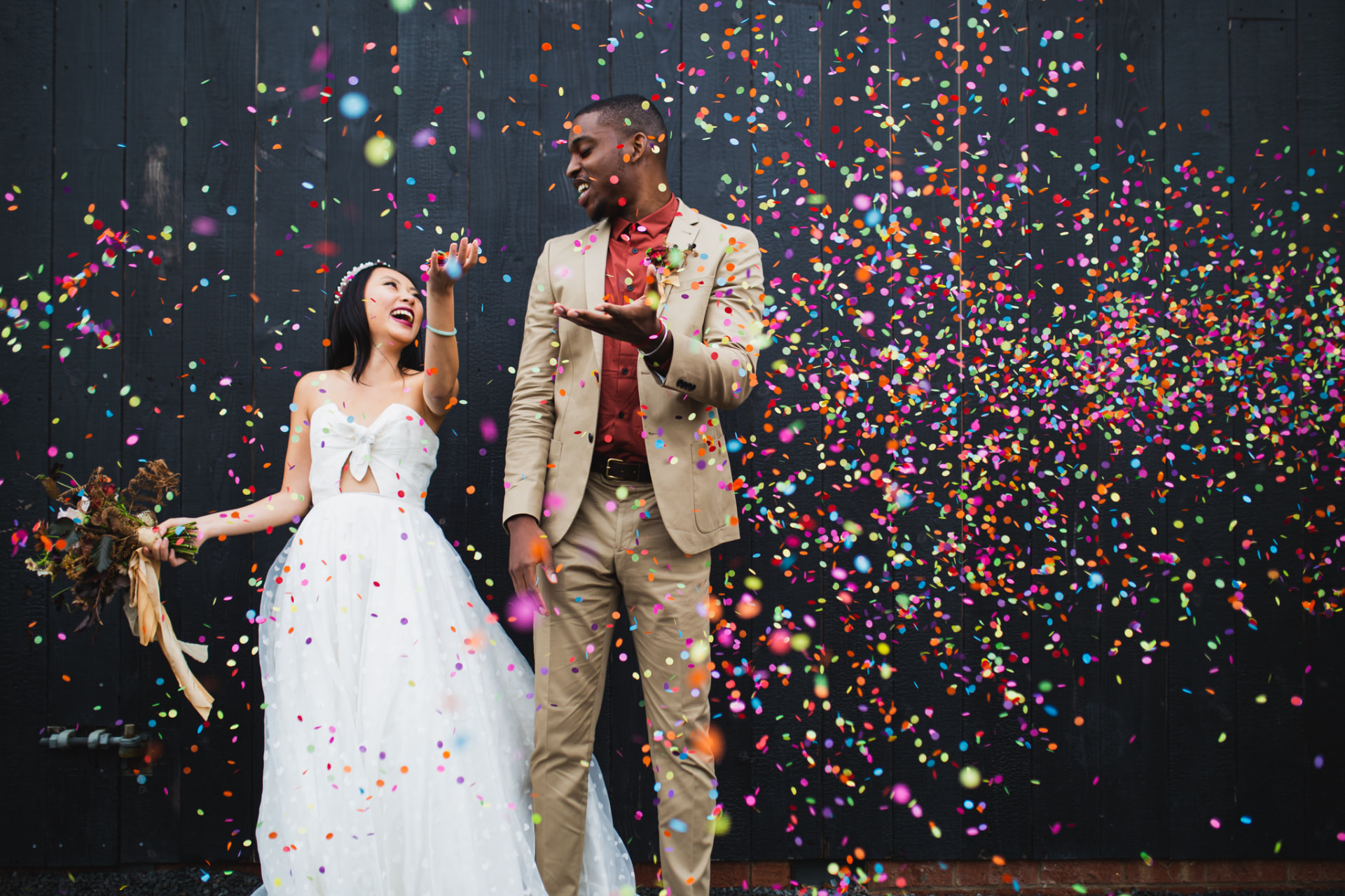 Newlyweds laughing beneath rainbow confetti against a dark wooden wall